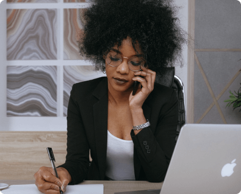 a woman with short, curly hair and glasses sitting at a desk with a laptop in front of her, talking on the phone and writing down something with a pen